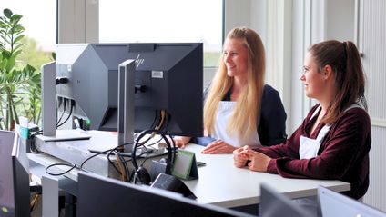 Zwei weibliche Führungskräfte aus der Qualitätssicherung bei Kaufland Fleischwaren stehen in einem Büro am Computer nebeneinander und arbeiten.