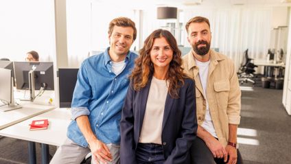 Großraumbüro: 3 Kaufland-Mitarbeiter posieren nebeneinander an der Kante eines Schreibtisches angelehnt für ein Gruppenfoto.