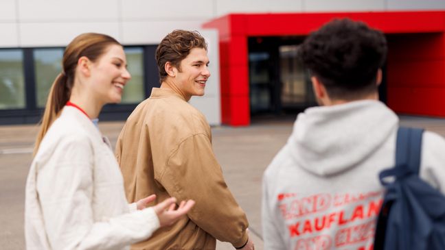 Drei Azubis gehen auf den Haupteingang der Kaufland-Zentrale zu. Links geht eine junge Frau, rechts neben ihr zwei junge Männer. Alle drei sind salopp gekleidet, man sieht sie in seitlicher Ansicht bzw. von hinten.