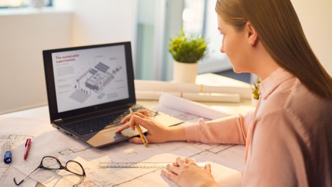 Eine Mitarbeiterin aus der Immobilienentwicklung betrachtet an ihrem Arbeitsplatz am Laptop den Entwurf eines Supermarkts.