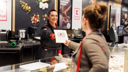 Eine Mitarbeiterin in scharzem Kaufland-Overall überreicht Kundin an der Bedientheke einen Käse.