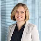 Frau mit blonden Haaren steht in schwarzer Bluse und beigem Blazer vor einer Glasfront und lächelt freundlich in die Kamera