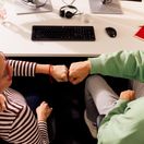 Kollege und Kollegin sitzen gemeinsam im Kaufland-Büro am Schreibtisch mit schwarzer Tastatur. Ihre zur Faust geballten Hände berühren sich zum Gruß.