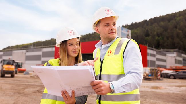 Zwei Mitarbeiter stehen vor dem Bau eines Kaufland-Marktes und schauen einen Bauplan an.