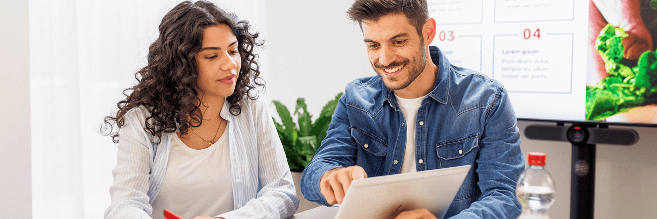 Eine weibliche Nachwuchskraft und ein Student in legerer Kleidung stehen vor einem aufgeklappten Laptop am Hochtisch. Der Student zeigt auf den nicht sichtbaren Bildschirm. Die beiden unterhalten sich.
