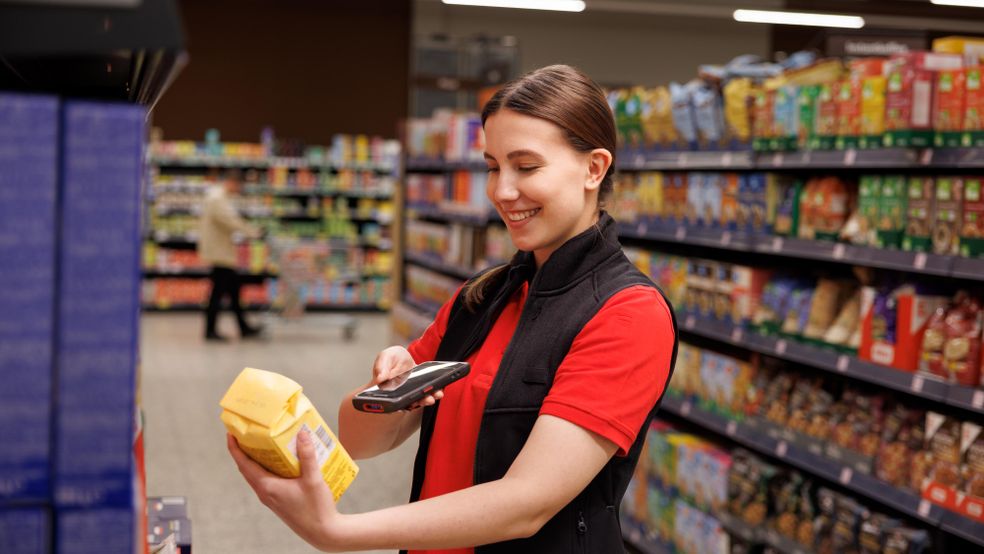 O femeie cu părul șaten strâns în coadă, zâmbitoare, îmbrăcată într-un tricou roșu și o vestă neagră, scanează codul de bare al unui pachet galben cu ajutorul unui dispozitiv mobil sau al unui scaner portabil într-un magazin alimentar.  În fundal se observă rafturi de supermarket pline cu diverse produse ambalate, iar în plan secund, o altă persoană este vizibilă estompat, mergând pe culoar.