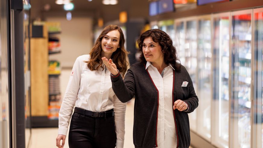 Eine weibliche Führungskraft und eine Mitarbeiterin befinden sich auf einem Rundgang durch eine Kaufland-Filiale. Die beiden sind im Gespräch. Hinter ihnen sind übermannsgroßen Kühlschränken zu sehen.