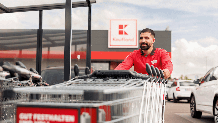 Ein Mitarbeiter in rot-schwarzer Dienstkleidung räumt außen vor einer Kaufland-Filiale Einkaufswagen auf.