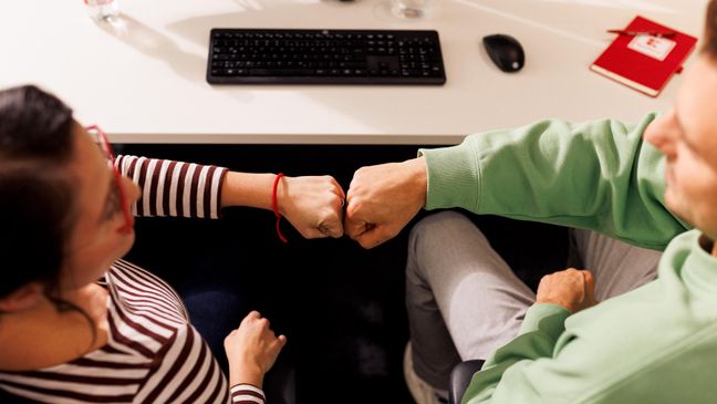 Kollege und Kollegin sitzen gemeinsam im Kaufland-Büro am Schreibtisch mit schwarzer Tastatur. Ihre zur Faust geballten Hände berühren sich zum Gruß.