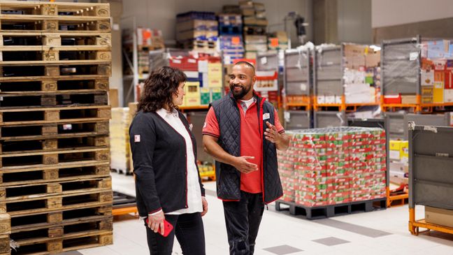 Im Filiallager: Kaufland-Verkaufsmitarbeiter im Gespräch mit einer Kollegin. Im Hintergrund ein Stapel Paletten.