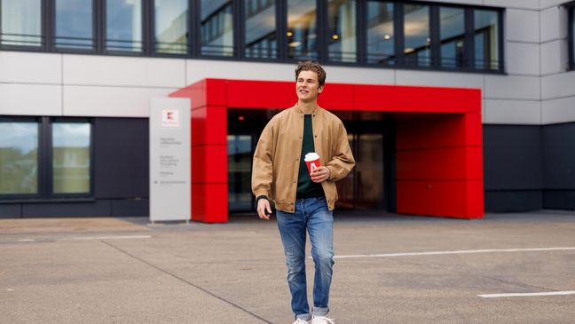 Ein Azubi, ein junger Mann in brauner Jacke, geht über den Platz vor dem Haupteingang der Kaufland-Zentrale.