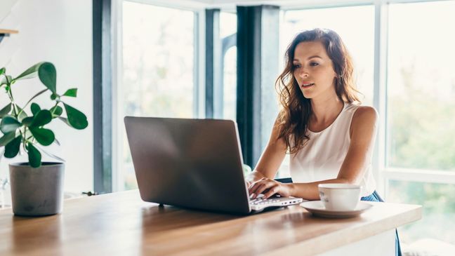 Frau sitzt daheim vor großen Fenstern und arbeitet an einem Laptop.