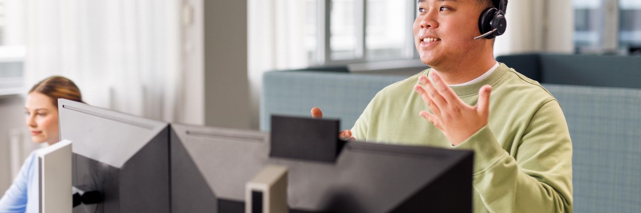 Ein Azubi sitzt mit Headset und im hellgrünen Pullover vor einem Computer. Er führt ein Gespräch.