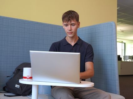 Dualer Student sitzt auf einem blauem Sofa und arbeitet konzentriert an einem Laptop.