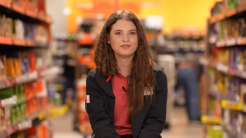 Portretul unei femei cu părul lung și ondulat, îmbrăcată într-o uniformă formată din tricou roșu și jachetă neagră, cu un ecuson în piept. Aceasta se află în centrul cadrului, pe culoarul unui magazin alimentar, având în fundal rafturi pline cu produse
