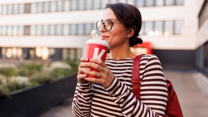 Mitarbeiterin in geringeltem Langarm-T-Shirt mit To-Go-Getränk vor einem Kaufland-Bürogebäude.