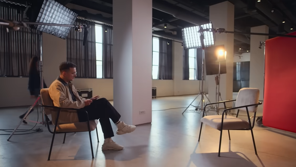 Un bărbat așezat pe un scaun într-un studio spațios de filmare sau fotografie, având în față un scaun gol și echipamente profesionale de iluminat.