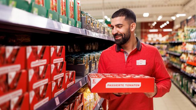 Ein Mitarbeiter in roter Kaufland-Kleidung steht in einem Gang, trägt einen Karton und verräumt Ware.