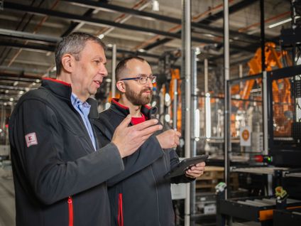 Teamleiter für Automatisierungstechnik erklärt einem Mitarbeiter etwas und zeigt mit der Hand auf eine Anlage.
