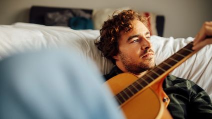 Mann sitzt auf dem Boden vor einem Bett und spielt Gitarre.
