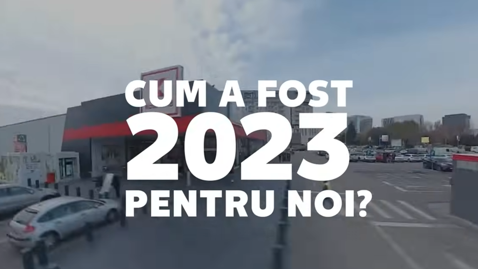 Iată descrierea detaliată a imaginii sub formă de text alternativ (alt text):  Text alternativ: O imagine exterioară, surprinsă dintr-un unghi ușor elevat, de tip dronă sau perspectivă largă, care înfățișează parcarea și fațada unui magazin Kaufland, recunoscut după sigla parțial vizibilă de pe acoperiș. În stânga se află clădirea modernă a hipermarketului, în nuanțe de gri închis și alb, cu o dungă roșie decorativă, iar în dreapta se întinde o parcare exterioară cu mai multe mașini. Peste această scenă este suprapus un text central de mari dimensiuni, scris cu majuscule albe: „CUM A FOST 2023 PENTRU NOI?”, unde anul „2023” este evidențiat printr-un font considerabil mai mare. Cerul este parțial acoperit de nori, sugerând o zi luminoasă de zi.