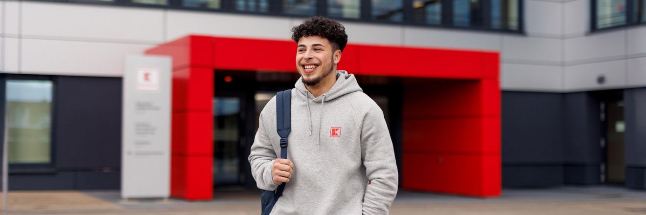 Ein Azubi, ein junger Mann in grauem Hoodie, geht über den Platz vor dem Haupteingang der Kaufland-Zentrale.