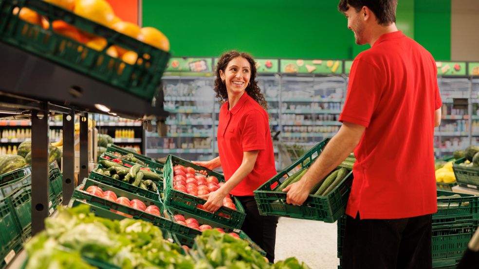 Doi angajați în uniforme roșii zâmbesc și colaborează în timp ce aranjează lădițe cu fructe și legume proaspete în raionul de supermarket.