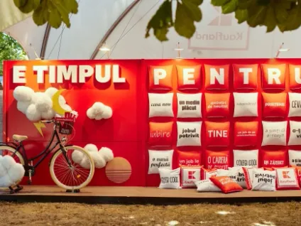 O imagine exterioară ce prezintă un stand publicitar de mari dimensiuni, amenajat pe o suprafață de pământ, sub o structură de tip cort alb. Fundalul standului este complet roșu și este împărțit în două secțiuni principale:  În partea stângă: Textul alb tridimensional „E TIMPUL” este poziționat deasupra unui decor fantezist. Acesta include o bicicletă neagră cu roți crem, decorată cu norișori albi și pufoși, o semilună galbenă și un fulger decorativ.  În partea dreaptă: Un panou format dintr-o matrice de perne pătrate, roșii și albe. Pernele de pe rândul de sus poartă literele albe care completează mesajul: „P E N T R U”. Celelalte perne au inscriși diverși termeni și mesaje în limba română, precum „ceva nou”, „un mediu plăcut”, „jobul potrivit”, „TINE”, „prietenie” sau „colegi cool”.  La baza panoului din dreapta se află o grămadă de perne decorative similare, unele purtând logo-ul Kaufland. Întregul stand este iluminat de reflectoare mici montate în partea de sus.