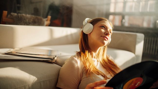 Frau sitzt vor einem Sofa und hört mit Kopfhörern Musik. In den Händen hält sie eine Schallplatte.