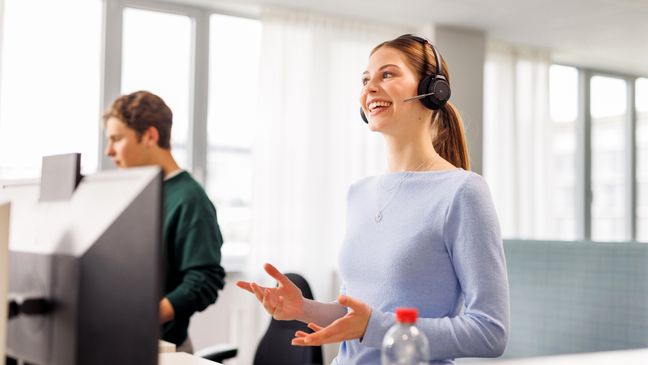 Ein weiblicher Azubi in hellblauem Pullover hat ein Headset auf und führt im Stehen ein Gespräch. Im Hintergrund sieht man halb verdeckt von einem Computer einen weiteren Azubi.