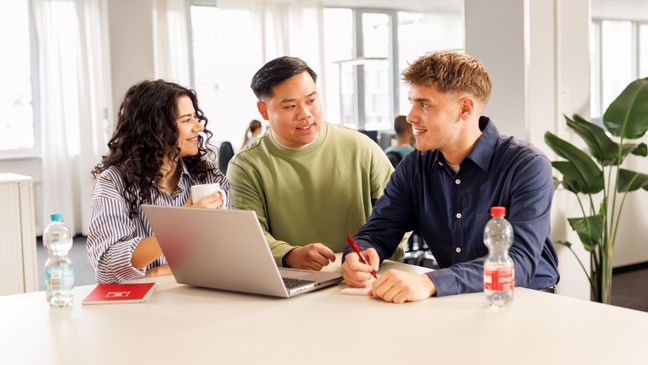 Kollaboratives Arbeiten mit einem Azubi, einem Mitarbeiter und einer Studentin. Die drei jungen Leute stehen in legerer Kleidung am Hochtisch vor einem aufgeklappten Laptop und unterhalten sich.