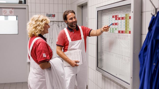 Eine Mitarbeiterin und ein Mitarbeiter in weißer Arbeitskleidung vor einem Schaukasten in einem Kaufland-Fleischwerk.