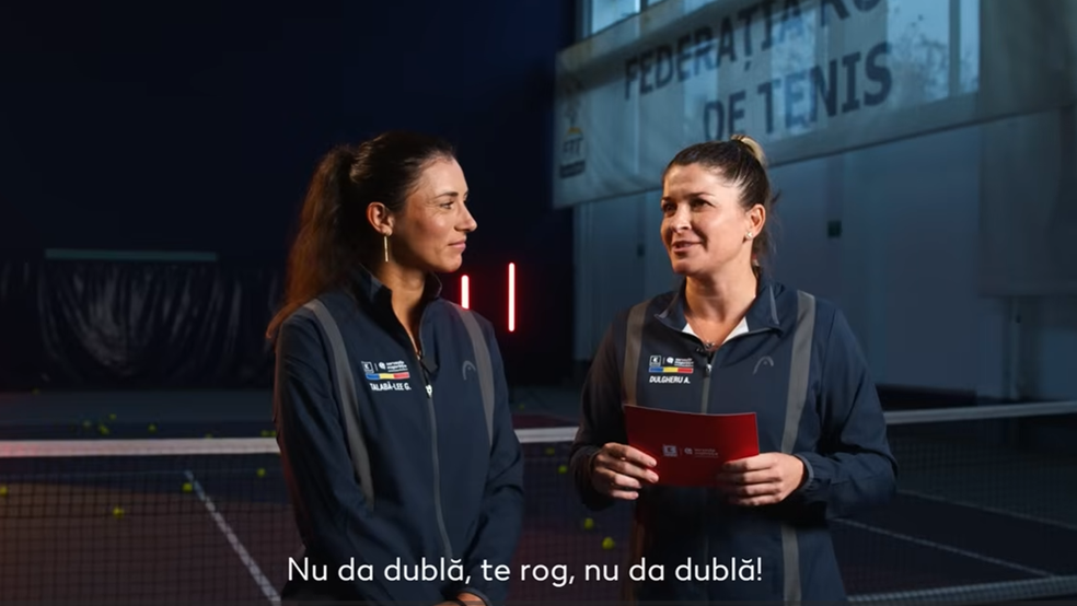 O imagine realizată în interiorul unei săli de tenis acoperite, înfățișând două femei într-un cadru de interviu. Ambele poartă jachete sport bleumarin identice, cu dungi gri pe umeri și logo-uri oficiale.  În stânga: O femeie cu părul închis la culoare prins în coadă de cal, având imprimat pe jachetă numele „TALABĂ-LEE G.” și steagul României. Ea privește zâmbitoare spre colega ei.  În dreapta: A doua femeie, cu părul prins într-un coc, are imprimat pe jachetă numele „DULGHERU A.”. Ea ține în mâini foi cartonate de culoare roșie și vorbește, privind spre stânga.  În fundal se observă fileul de tenis, mingi galbene pe teren și un banner alb mare pe care scrie parțial „FEDERAȚIA ROMÂNĂ DE TENIS”. În partea de jos a imaginii este adăugat un subtitlu alb: „Nu da dublă, te rog, nu da dublă!”.