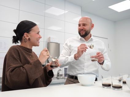 Zwei Mitarbeiter bei Kaufland International verkosten gemeinsam Kaffee und lächeln sich an.
