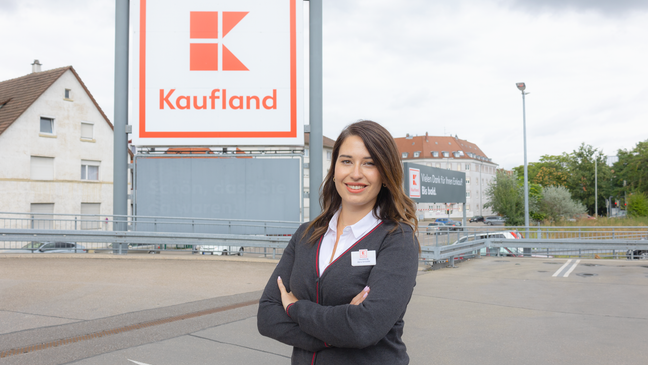 Hausleiterin und ehemalige Trainee steht vor Kaufland-Filiale und lächelt in die Kamera.
