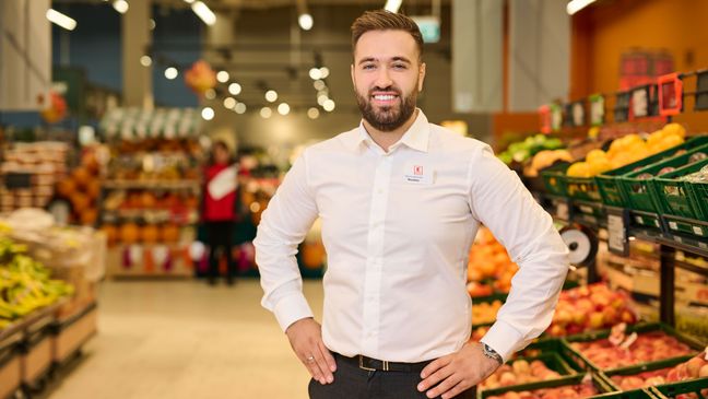 Ein lächelnder Kaufland-Hausleiter in weißem Hemd steht selbstbewusst mit in die Hüften gestemmten Armen in der Obst- und Gemüseabteilung einer modernen Kaufland-Filiale; Karrieremöglichkeiten im Einzelhandel.