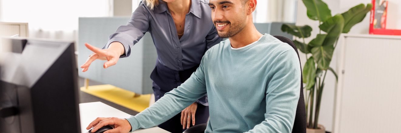 Eine weibliche Führungskraft erläutert einem Trainee in türkisfarbenem Longsleeve etwas. Der Trainee sitzt vor einem Computer, die Frau steht links hinter ihm und zeigt auf den Bildschirm.