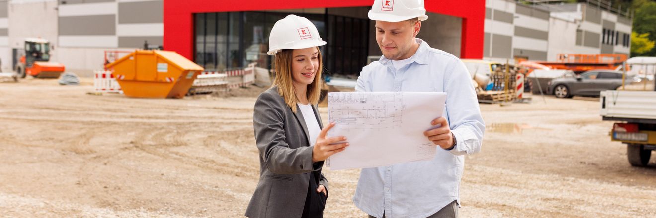 Zwei Mitarbeiter stehen vor dem Bau eines Kaufland-Marktes und schauen einen Bauplan an.