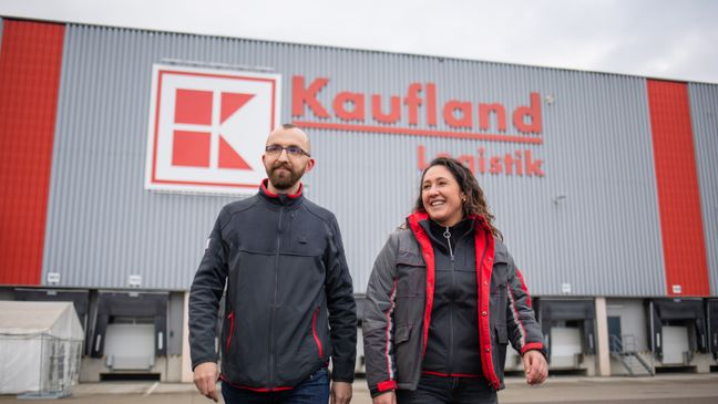 Zwei Logistik-Mitarbeiter bei Kaufland laufen vor einem Logistikzentrum und lachen.