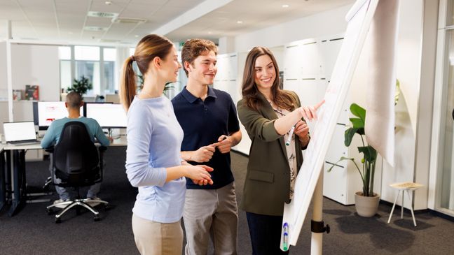 Zwei Azubis, eine junge Frau und ein junger Mann, sowie eine Ausbilderin befinden sich in einer Workshopsituation. Die drei stehen vor einem Flipchart und brainstormen.