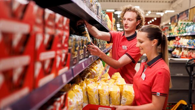 Regalpflege: Ein Schüler und eine Schülerin in roter Kaufland-Kleidung räumen Ware in Regale ein.