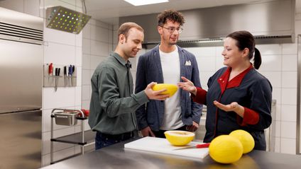 Zwei Mitarbeiter und eine Mitarbeiterin stehen in einer Laborumgebung am Tisch. Sie nehmen eine Verkostung vor und prüfen Ware.