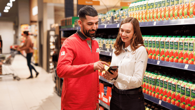 Ein Mitarbeiter in roter Kaufland-Kleidung und eine Mitarbeiterin mit weißem Hemd stehen in einem Gang und tauschen sich aus.