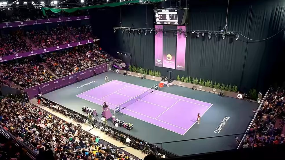 Iată descrierea detaliată a imaginii sub formă de text alternativ (alt text):  Text alternativ: O imagine panoramică, realizată de la înălțime, care surprinde atmosfera dintr-o arenă sportivă interioară plină de spectatori în timpul unui meci de tenis. În centrul arenei se află terenul de tenis cu suprafață mov aprins și margini gri închis, pe care sunt poziționate cele două jucătoare de o parte și de alta a fileului. Pe marginile terenului este scris textul „CLUJ-NAPOCA”.  Tribunele din partea stângă și din fundal sunt pline de public, iar pe ecranele și bannerele din arenă se disting lumini și elemente de branding în nuanțe de roz și violet. Structura tehnică de lumini este suspendată deasupra terenului, iar în partea de sus, central, este vizibil tabela electronică de marcaj.