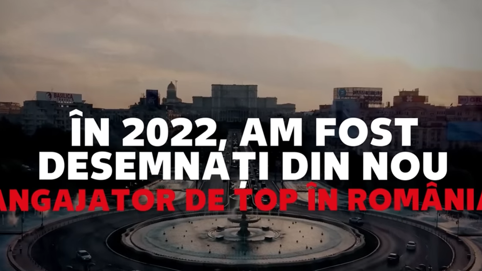 O imagine panoramică de la înălțime care surprinde Piața Unirii din București în amurg, având în fundal Palatul Parlamentului. În prim-plan se află fântânile arteziene și un sens giratoriu cu trafic auto redus. Peste imagine este suprapus un text mare, scris cu majuscule. Rândurile de sus sunt albe și transmit mesajul: „ÎN 2022, AM FOST DESEMNAȚI DIN NOU”, iar rândul de jos este roșu și completează fraza: „ANGAJATOR DE TOP ÎN ROMÂNIA”. Pe clădirile din fundal se pot observa câteva reclame luminoase