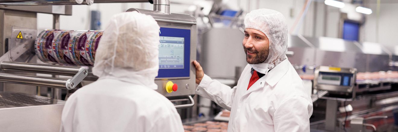 Fertigungsanlage in einem Kaufland-Fleischwerk. Zwei Kaufland-Mitarbeiter bedienen eine Produktionsanlage und unterhalten sich.