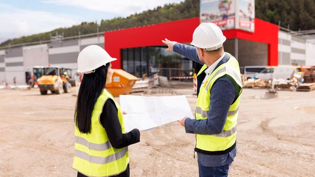 Zwei Mitarbeiter stehen vor dem Bau eines Kaufland-Marktes und sprechen über einen Bauplan, den sie in den Händen halten. Der Mitarbeiter rechts zeigt auf den Bau.