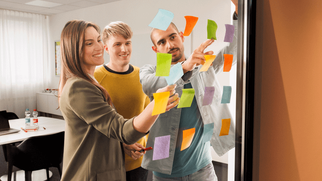 Eine junge Frau, ein junger Mann, beide Trainees und ein Ausbilder sind in einem Meeting. Die drei stehen vor einer gläsernen Wand und arbeiten mit Post-Its.