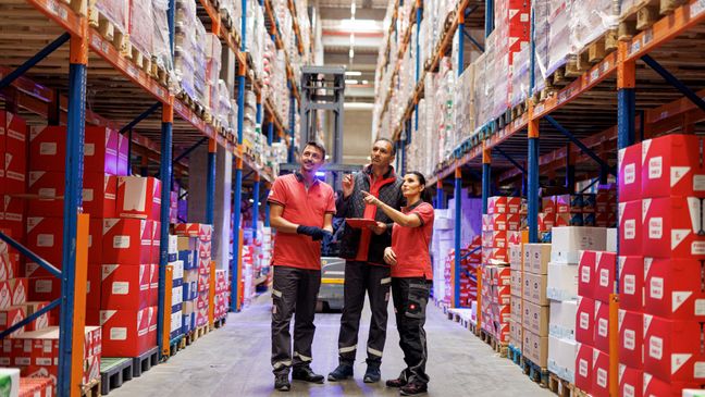 3 Logistikmitarbeiter von Kaufland in einem Lagergang mit hohen, gut gefüllten Regalen.