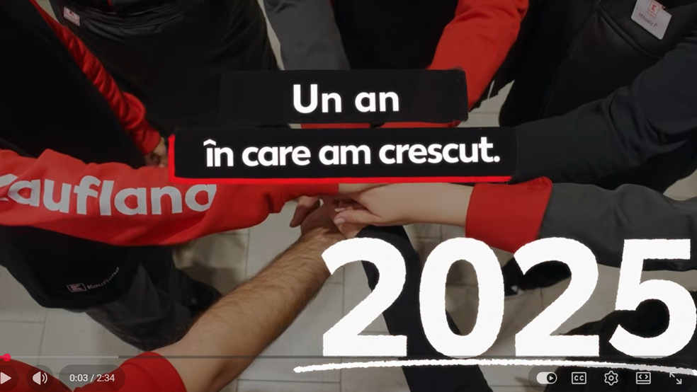 O imagine filmată de sus ce surprinde un grup de angajați care își pun mâinile una peste alta în centru, sugerând un gest de echipă și solidaritate. Persoanele poartă echipament de lucru în culorile roșu, negru și gri; pe o mânecă roșie se observă clar logo-ul alb „Kaufland”, iar în colțul din dreapta sus apare un ecuson pe o vestă. În centrul imaginii este suprapus un text alb pe fundal negru: „Un an în care am crescut.”, iar în colțul din dreapta jos este scris, cu un font mare alb ce imitătul scrisul cu cretă, anul „2025”. În partea inferioară se pot observa elemente specifice unui player video (bara de redare și butoanele de control).